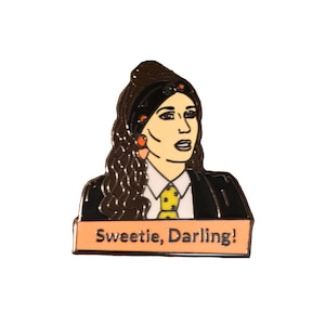 Könnte beinhalten: Emaille-Anstecknadel mit einer stilisierten Illustration einer Frau mit langen braunen Haaren, die einen schwarzen Blazer, ein weißes Hemd und eine gelbe Krawatte mit grünen Punkten trägt. Die Nadel enthält den Text "Sweetie, Darling!" auf einem pfirsichfarbenen Banner.
