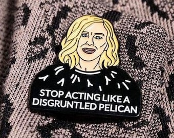 Può includere: Spilla in smalto nero con l'illustrazione di una donna bionda. La spilla presenta la scritta bianca "STOP ACTING LIKE A DISGRUNTLED PELICAN". La spilla è posta su un tessuto fantasia.