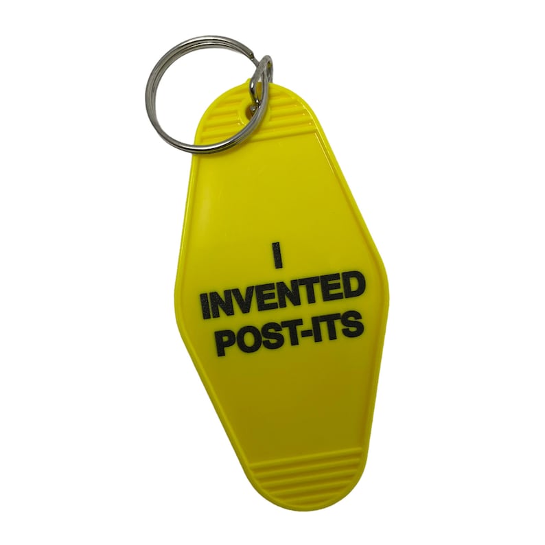 Pu&ograve; includere: Portachiavi in plastica giallo a forma di chiave di motel con il testo "Ho inventato i Post-it" in nero.