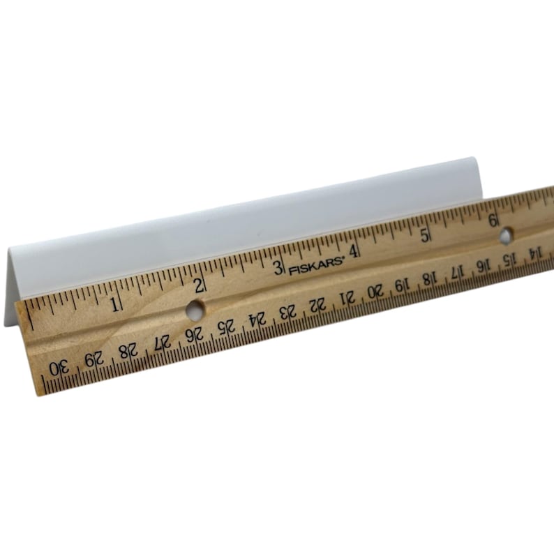 May include: A white, angled trim piece is positioned next to a wooden ruler. The ruler, marked with black increments, measures up to 30 inches. The trim piece appears to be made of a solid, opaque material.
