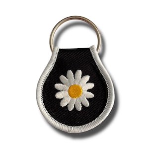 May include: Black keychain with a white daisy flower design. The daisy has a yellow center and white petals. The keychain has a silver metal ring and a white stitched border.