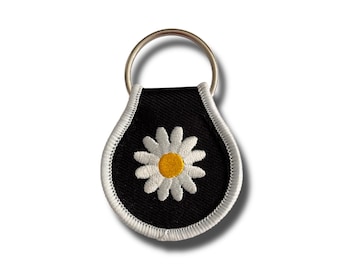 May include: Black keychain featuring a white daisy flower design with a yellow centre and white petals. The keychain has a silver metal ring and a white stitched border.