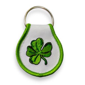 Peut inclure: Un porte-clés blanc avec un trèfle à quatre feuilles vert brodé dessus. Le porte-clés a un anneau en métal argenté.