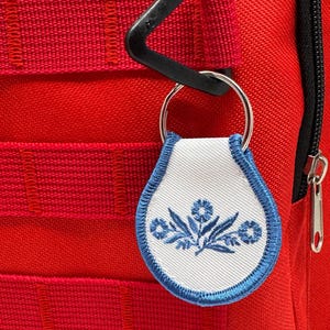 May include: A white fabric keychain with a blue embroidered floral design attached to a silver key ring. The keychain is attached to a red bag with black zipper pulls.