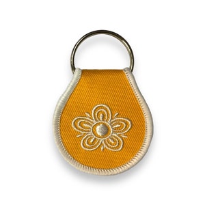 May include: A yellow key chain with a white embroidered flower design. The key chain has a silver metal ring.