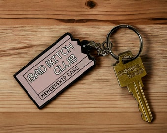 Può includere: Portachiavi rosa con bordo nero e il testo "Bad Bitch Club Membership Card". Il portachiavi è attaccato a un portachiavi argentato e una chiave dorata con il testo "minute key co."