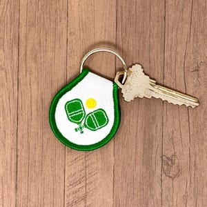 May include: A white and green keychain with a pickleball design. The keychain is attached to a silver key.
