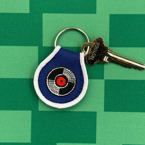 May include: A navy blue vinyl record keyring with a white border and a silver key ring. The record design features a black and white record with a red centre. A silver key is attached.