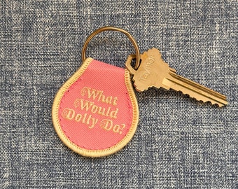 May include: A pink fabric keychain with the text "What Would Dolly Do?" in gold embroidery. The keychain is attached to a gold key.