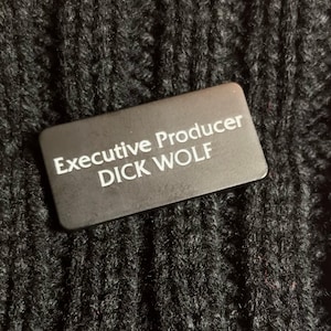 May include: Black rectangular name tag with white text that reads "Executive Producer DICK WOLF".