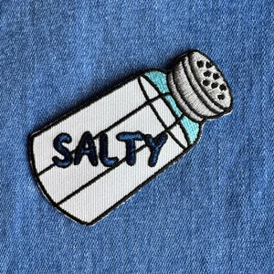 May include: White salt shaker with blue accents and the word "SALTY" in black lettering. The shaker is on a blue denim background.