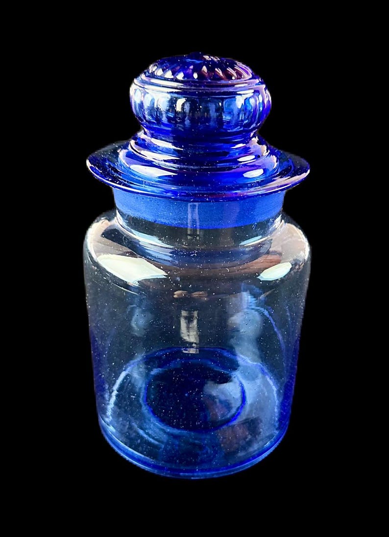 Peut inclure: Un pot de pharmacie en verre bleu cobalt avec un couvercle assorti. Le pot est cylindrique avec une base arrondie et un bord &eacute;vas&eacute;. Le couvercle est de forme arrondie avec un bouton sur le dessus. Le pot est vide.