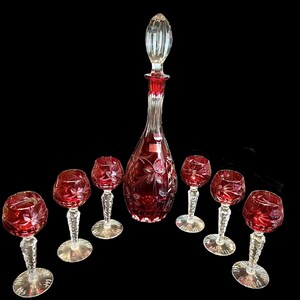 May include: A ruby red crystal wine decanter with a matching stopper and six wine glasses. The decanter and glasses feature a floral design. The set is made of clear crystal with red coloring.