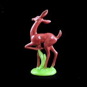 May include: A red ceramic deer figurine standing on a green base. The deer is posed with its head turned to the side and its front leg raised.