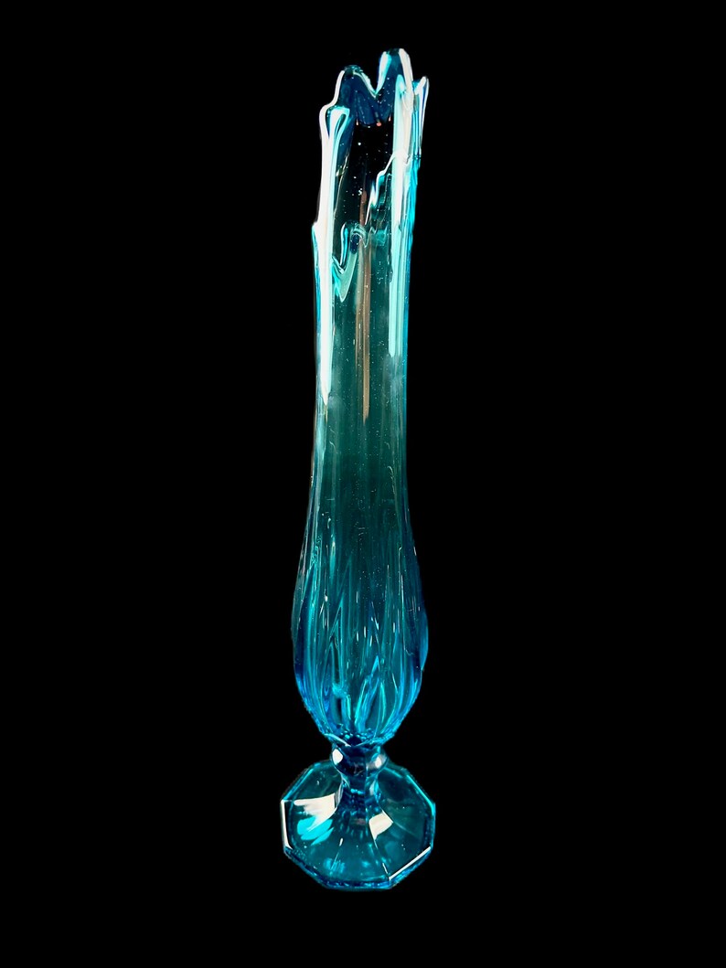 Pode incluir: Um vaso de vidro azul alto com uma base octogonal alargada e uma parte superior ondulada e texturizada.