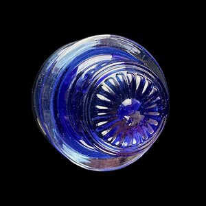 Peut inclure: Objet en verre transparent avec un int&eacute;rieur bleu profond. La base pr&eacute;sente un motif circulaire et stri&eacute; avec une indentation centrale arrondie. L'objet est pos&eacute; sur un fond noir, soulignant ses qualit&eacute;s transparentes et r&eacute;fl&eacute;chissantes.