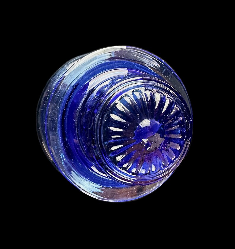 Peut inclure: Objet en verre transparent avec un int&eacute;rieur bleu profond. La base pr&eacute;sente un motif circulaire et stri&eacute; avec une indentation centrale arrondie. L'objet est pos&eacute; sur un fond noir, soulignant ses qualit&eacute;s transparentes et r&eacute;fl&eacute;chissantes.