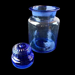 Peut inclure: Un bocal d'apothicaire en verre bleu clair avec un couvercle assorti. Le bocal a une large ouverture et un corps arrondi. Le couvercle est de forme plus petite et arrondie avec un motif d&eacute;coratif &agrave; nervures. Le bocal et le couvercle sont pos&eacute;s sur un fond noir.
