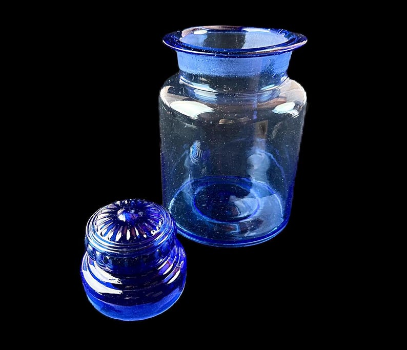 Peut inclure: Un bocal d'apothicaire en verre bleu clair avec un couvercle assorti. Le bocal a une large ouverture et un corps arrondi. Le couvercle est de forme plus petite et arrondie avec un motif d&eacute;coratif &agrave; nervures. Le bocal et le couvercle sont pos&eacute;s sur un fond noir.