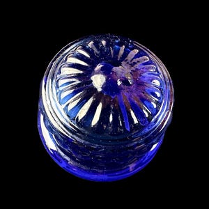 Peut inclure: Un pot en verre bleu cobalt avec un couvercle d&eacute;coratif. Le couvercle pr&eacute;sente un motif radial avec des accents blancs. Le pot est pos&eacute; sur un fond noir, soulignant sa couleur et sa forme.