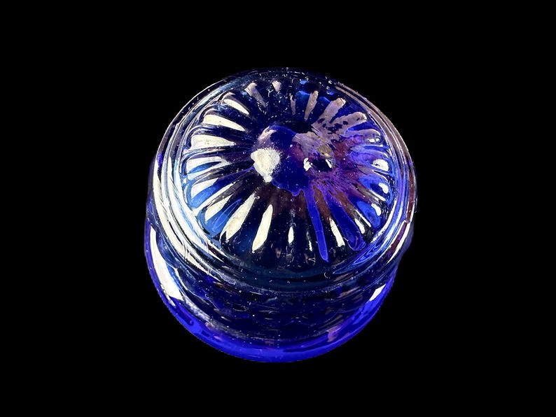 Peut inclure: Un pot en verre bleu cobalt avec un couvercle d&eacute;coratif. Le couvercle pr&eacute;sente un motif radial avec des accents blancs. Le pot est pos&eacute; sur un fond noir, soulignant sa couleur et sa forme.