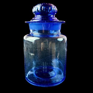 Peut inclure: Un pot d'apothicaire en verre bleu cobalt avec un couvercle assorti. Le pot est cylindrique avec une base arrondie et un bord &eacute;vas&eacute;. Le couvercle est de forme arrondie avec un bouton sur le dessus. Le pot est vide et se trouve sur un fond noir.