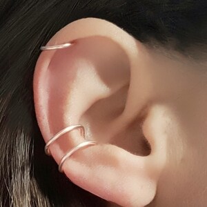 May include: Two thin gold ear cuffs worn on the upper cartilage of the ear.