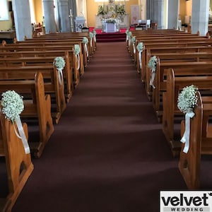 Puede incluir: Vista interior de una iglesia decorada para una boda. Bancos de madera bordean un pasillo alfombrado de rojo. Arreglos florales blancos con cintas adornan los bancos. Una vidriera se ve al final del pasillo.