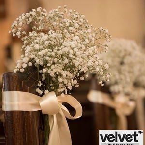 Puede incluir: Un ramo de flores blancas de paniculata atado a una silla de madera con una cinta de satén color crema. Las flores están en plena floración y la cinta está atada en un gran lazo. La imagen es para una boda.