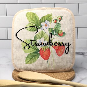 May include: A white cotton pot holder with a strawberry design. The pot holder features a watercolor illustration of strawberries, leaves, and flowers. The text "Strawberry" is written in black cursive.