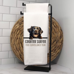 May include: White kitchen towel with a black Labrador Retriever peeking over the top. The towel has the text "Counter Surfer Food Surveillance Team" printed on it.
