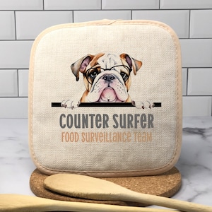 May include: A beige kitchen pot holder with a brown and white bulldog wearing glasses peeking over the top. The text "Counter Surfer Food Surveillance Team" is printed below the dog.