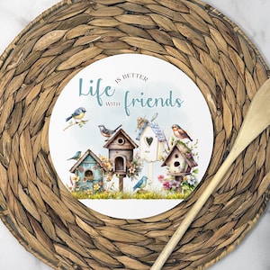 May include: A round decorative plate with a watercolor illustration of birdhouses and birds, with the text "Life is better with friends." The plate is set on a woven placemat with a wooden spoon.