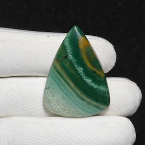 May include: A polished, triangular gemstone with a mix of green, yellow, and blue-green hues. The stone features a layered, banded pattern, with a smooth, glossy surface. The gemstone is held in a white glove against a black background.
