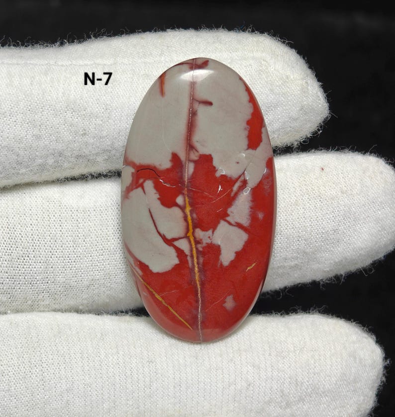 May include: An oval gemstone featuring a striking red and grey pattern, bisected by a thin, golden-yellow vein. The text "N-7" is visible in the upper left corner. The gemstone is displayed against a white fabric background.
