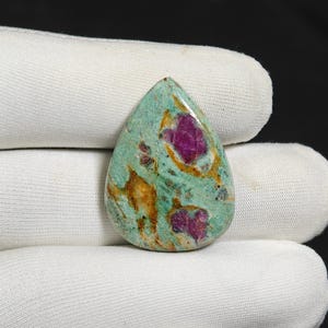 Puede incluir: Una gema en forma de lágrima con una base turquesa, con inclusiones magenta y marrón dorado vibrantes. La piedra pulida se encuentra sobre un fondo de tela blanca, mostrando su belleza natural y patrones únicos.