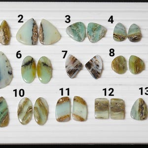 May include: Thirteen pairs of green, brown, and white opal cabochons, numbered 1 through 13. The cabochons are arranged in two rows on a white surface.