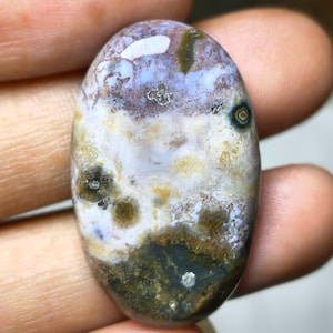 May include: An oval-shaped gemstone with a unique pattern of colors. The stone features a mix of white, light blue, brown, and dark green hues. The polished surface reflects light, highlighting the natural variations and textures within the stone.