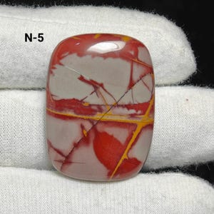May include: A polished, rectangular gemstone with red, grey, and yellow patterns. The stone has a smooth, glossy surface and is held by a white cloth. The text "N-5" is visible in the upper left corner.
