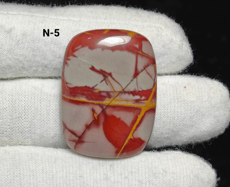 May include: A polished, rectangular gemstone with red, grey, and yellow patterns. The stone has a smooth, glossy surface and is held by a white cloth. The text "N-5" is visible in the upper left corner.