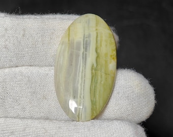 May include: An oval-shaped gemstone with a polished surface. The stone features a blend of pale yellow, green, and gray hues, with vertical striations creating a textured appearance. The gemstone is held against a white fabric background.