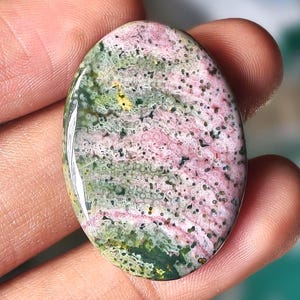 May include: An oval-shaped gemstone with a polished surface. The stone features a mix of colors, including pink, green, and black speckles. The gemstone is held in a hand, showcasing its size and texture. The overall appearance suggests a natural, handcrafted item.