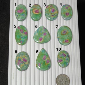 Könnte beinhalten: Eine Sammlung von zehn polierten Edelsteinen in verschiedenen Formen, darunter oval und tropfenförmig. Die Steine haben eine grüne Grundfarbe mit rosa und gelben Einschlüssen. Eine US-Viertel-Münze dient als Größenvergleich.
