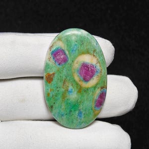 Puede incluir: Gema ovalada con base verde, con inclusiones rosas y rojas. La piedra pulida se muestra sobre un fondo de tela blanca. Las inclusiones están rodeadas por un halo color crema, creando un contraste llamativo.