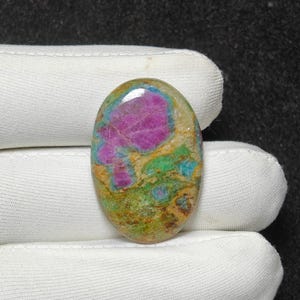 Könnte beinhalten: Oval geformter Edelstein mit einer lebendigen Farbmischung. Der Stein weist eine markante magenta Mitte auf, die von Bereichen in Grün, Blau und Beige umgeben ist. Die polierte Oberfläche reflektiert das Licht und hebt die natürlichen Muster hervor.