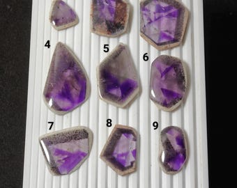 May include: Nine polished amethyst gemstones of varying shapes and sizes are displayed on a white surface. The gemstones showcase a range of purple hues with unique internal patterns and textures. A coin is included for size reference.