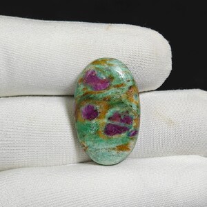 Puede incluir: Gema ovalada con superficie pulida. La piedra presenta una mezcla de colores, incluyendo verde, marrón y parches de rojo intenso. La gema está sobre un fondo de tela blanca.