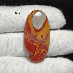 May include: An oval gemstone with a polished surface. The stone displays a mix of red, yellow, and grey colours, with detailed patterns and veining. The text "N-2" is visible on the top left. The gemstone is held against a white fabric background.