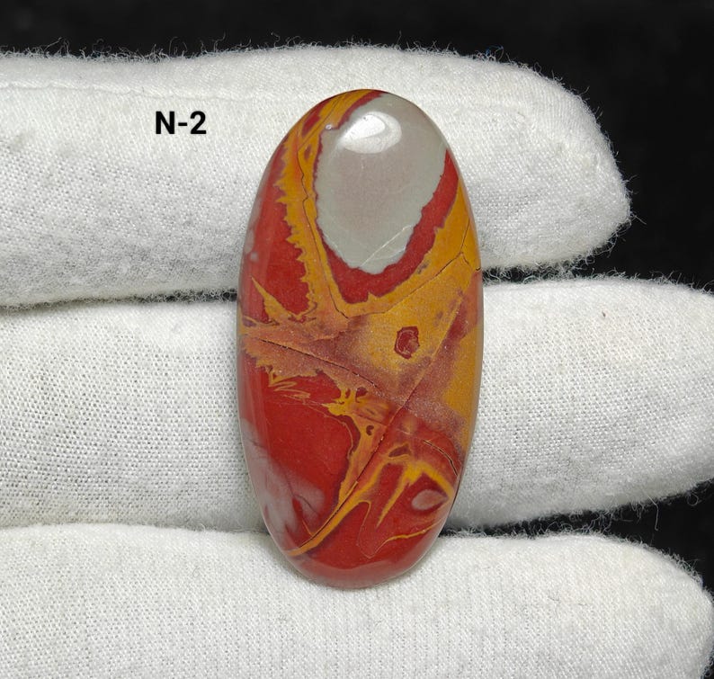 May include: An oval gemstone with a polished surface. The stone displays a mix of red, yellow, and grey colours, with detailed patterns and veining. The text "N-2" is visible on the top left. The gemstone is held against a white fabric background.