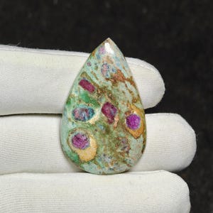 Puede incluir: Una piedra preciosa en forma de lágrima con una matriz moteada en tonos verdes y bronce, que presenta inclusiones de rubí en color magenta vibrante. La superficie pulida muestra una textura suave y redondeada con variaciones de color.